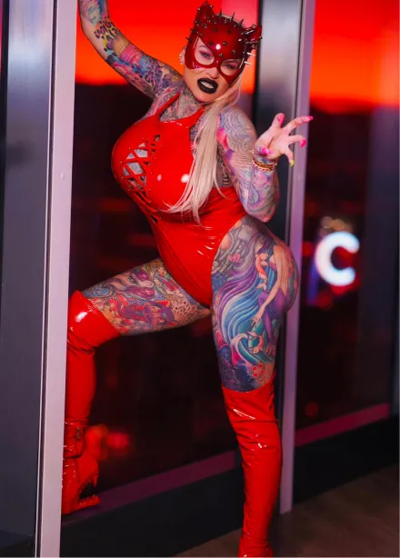 Person wearing a red latex bodysuit and matching thigh-high boots with elaborate tattoos covering their arms and legs, posing against a window with a red sunset background.