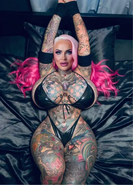 Woman with long pink curly pigtails, glasses, and pink leopard print top and underwear, posing against a solid pink background with visible tattoos on her torso and legs.