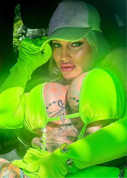 Woman with bright green hair, wearing a matching green hat with a pink flower, a sequined green bikini top, and displaying colorful tattoos on her arms and chest, posing outdoors on green grass.