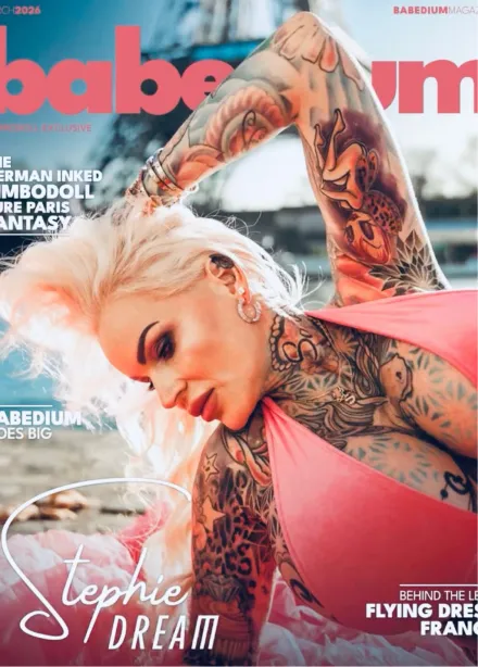 Blonde woman with extensive colorful tattoos wearing a coral dress, posing outdoors with the Eiffel Tower blurred in the background.