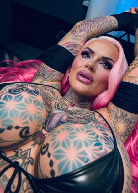 Blonde woman with tattoos wearing a tight pink dress standing on a sunny street with palm trees and a pink car behind her.