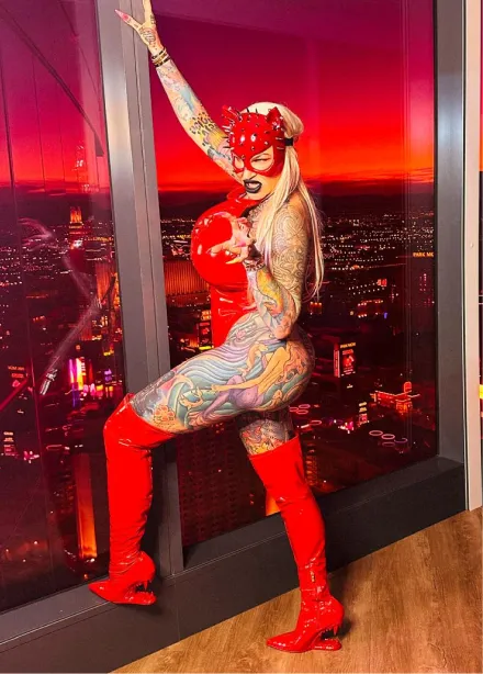 Blonde woman with tattoos wearing a tight pink dress standing on a sunny street with palm trees and a pink car behind her.