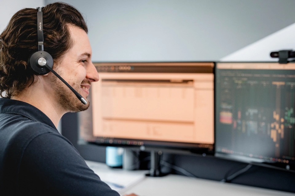 Ein Mann mit Headset lächelt und arbeitet vor zwei Computermonitoren in einem Büro.