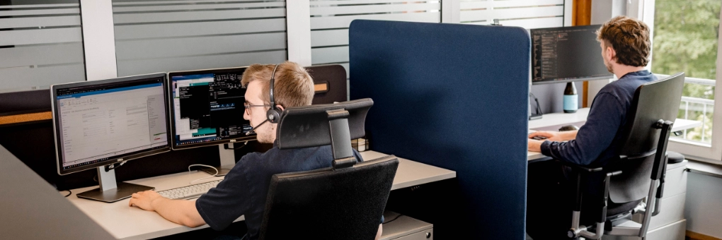 Zwei Männer arbeiten an Büroarbeitsplätzen mit Computern und Trennwänden, einer mit Headset.