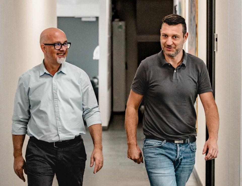 Zwei Männer, einer mit Brille und Hemd, der andere in Poloshirt und Jeans, gehen und unterhalten sich im Flur.