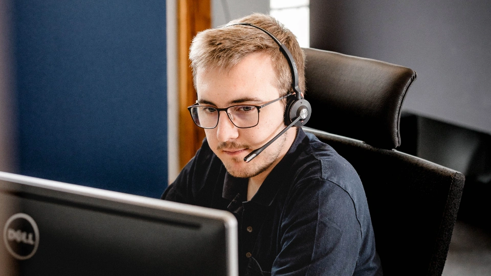 Junger Mann mit Brille und Headset arbeitet konzentriert an einem Computerbildschirm.