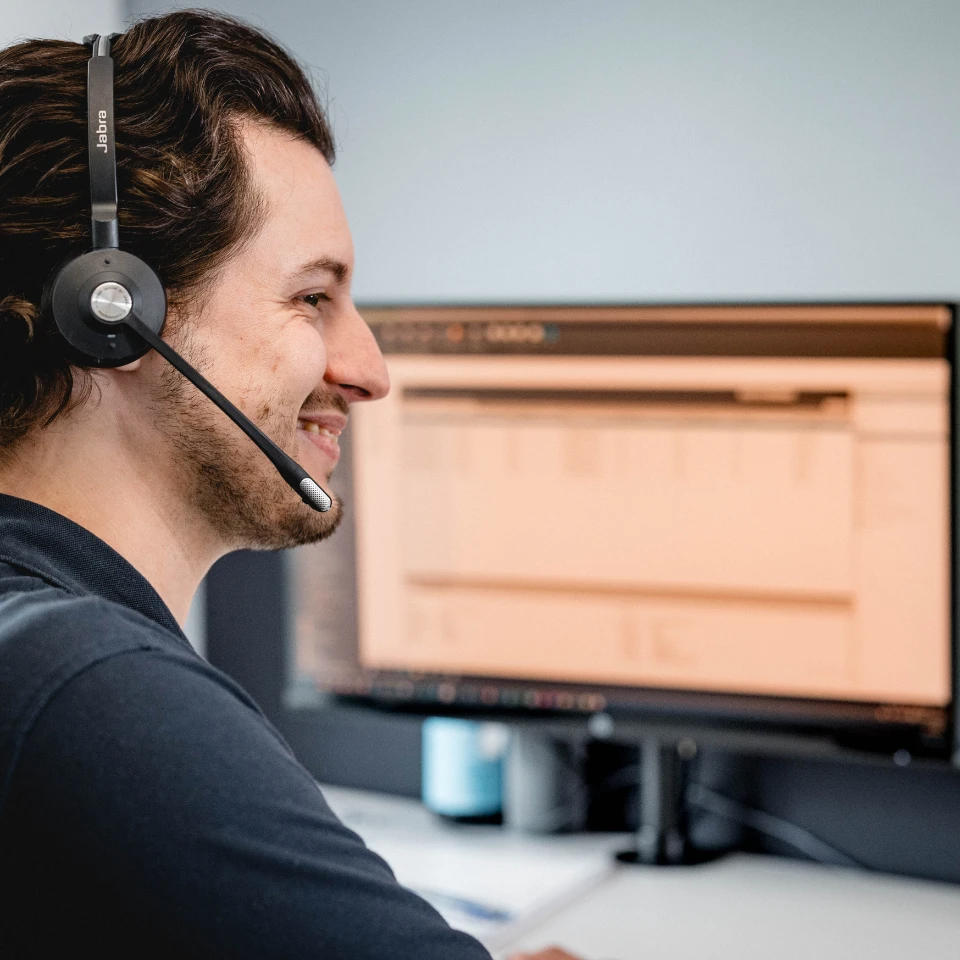 Mann mit Headset lächelt vor einem großen Computermonitor mit geöffneter Software.