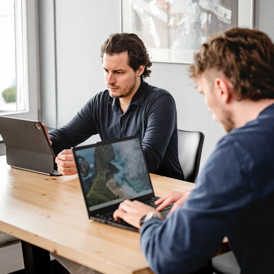 Zwei Männer sitzen am Tisch und arbeiten konzentriert an Tablet und Laptop in einem hellen Raum.