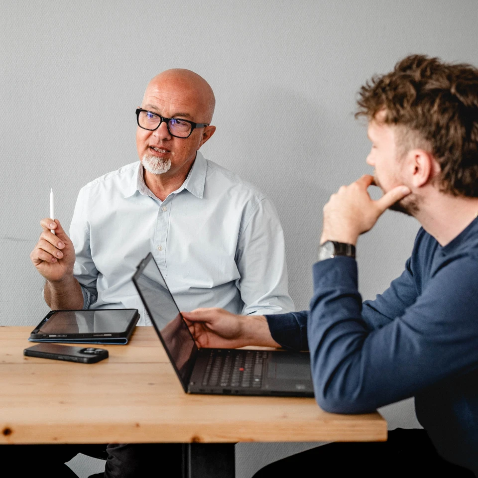 Zwei Männer im Gespräch an einem Tisch mit Laptop, Tablet und Smartphone.