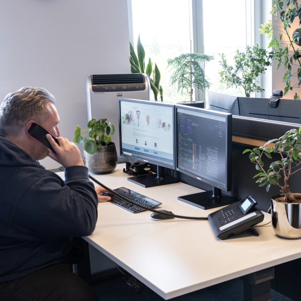 Mann am Schreibtisch telefoniert mit zwei Monitoren und Pflanzen in einem modernen Büro.