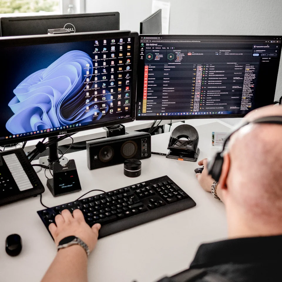 Person am Schreibtisch mit zwei Monitoren, Tastatur, Maus und Headset, die IT-Software überwachen.