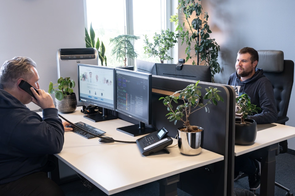 Zwei Männer arbeiten an gegenüberliegenden Schreibtischen in einem modernen Büro mit mehreren Monitoren und Pflanzen.