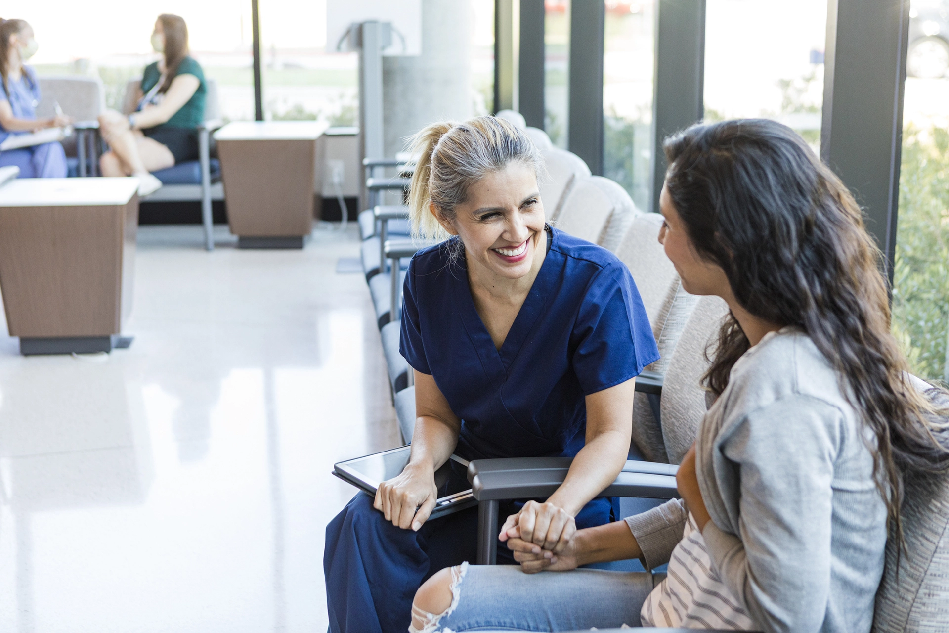 A smiling female nurse in blue scrubs holding hands and talking with a seated woman in a waiting room.