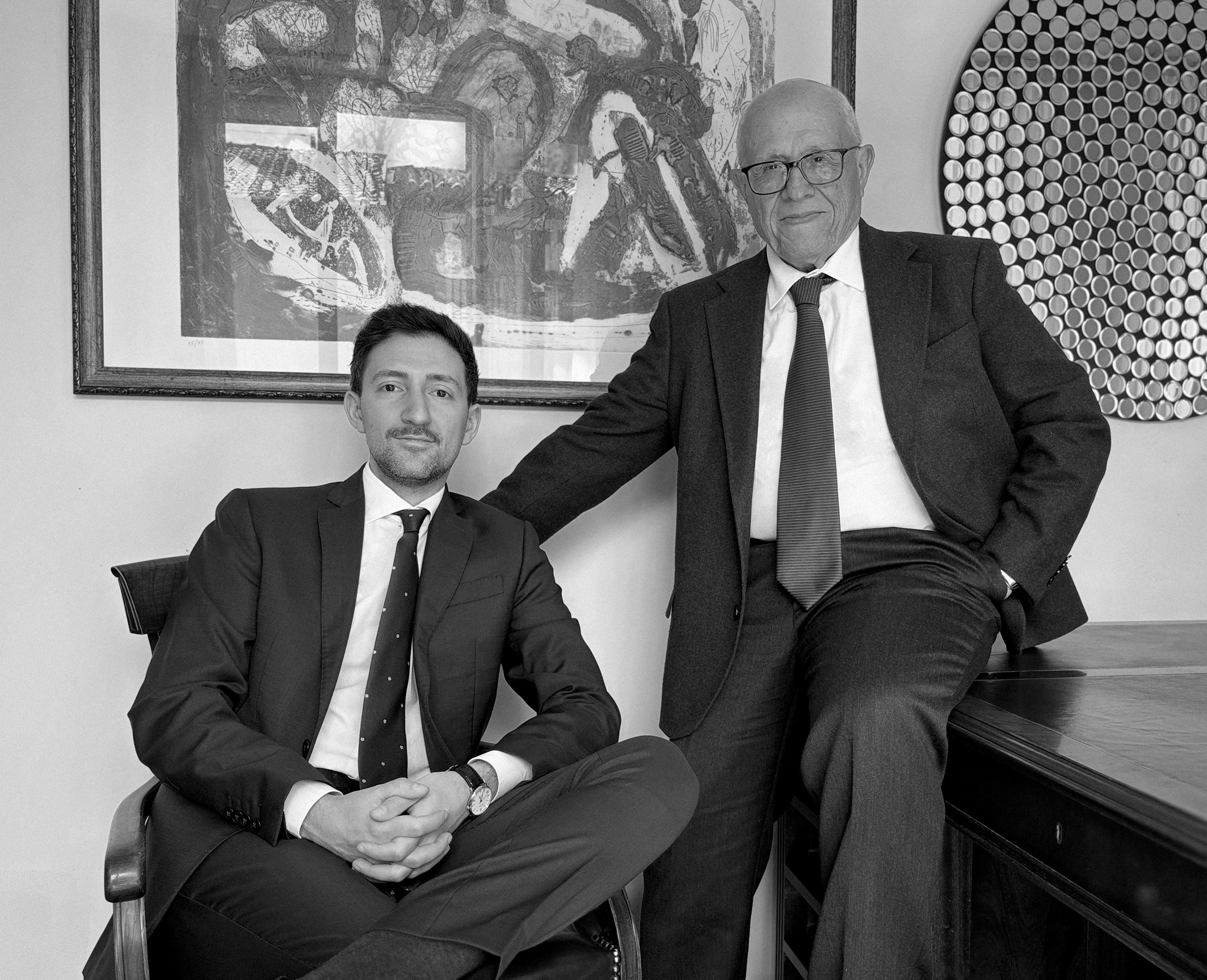 Due uomini in abiti formali, uno seduto e l'altro appoggiato a una scrivania, con un quadro astratto sullo sfondo.