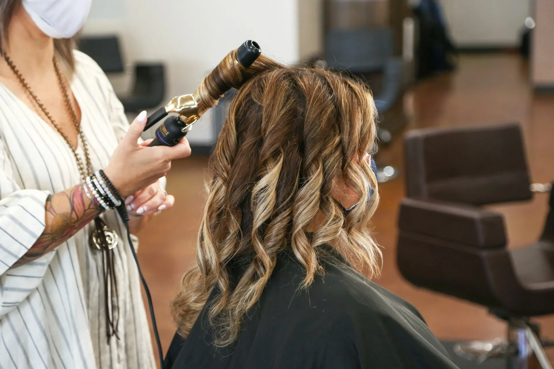 Hairdresser curling a woman's shoulder-length, highlighted hair in a salon.