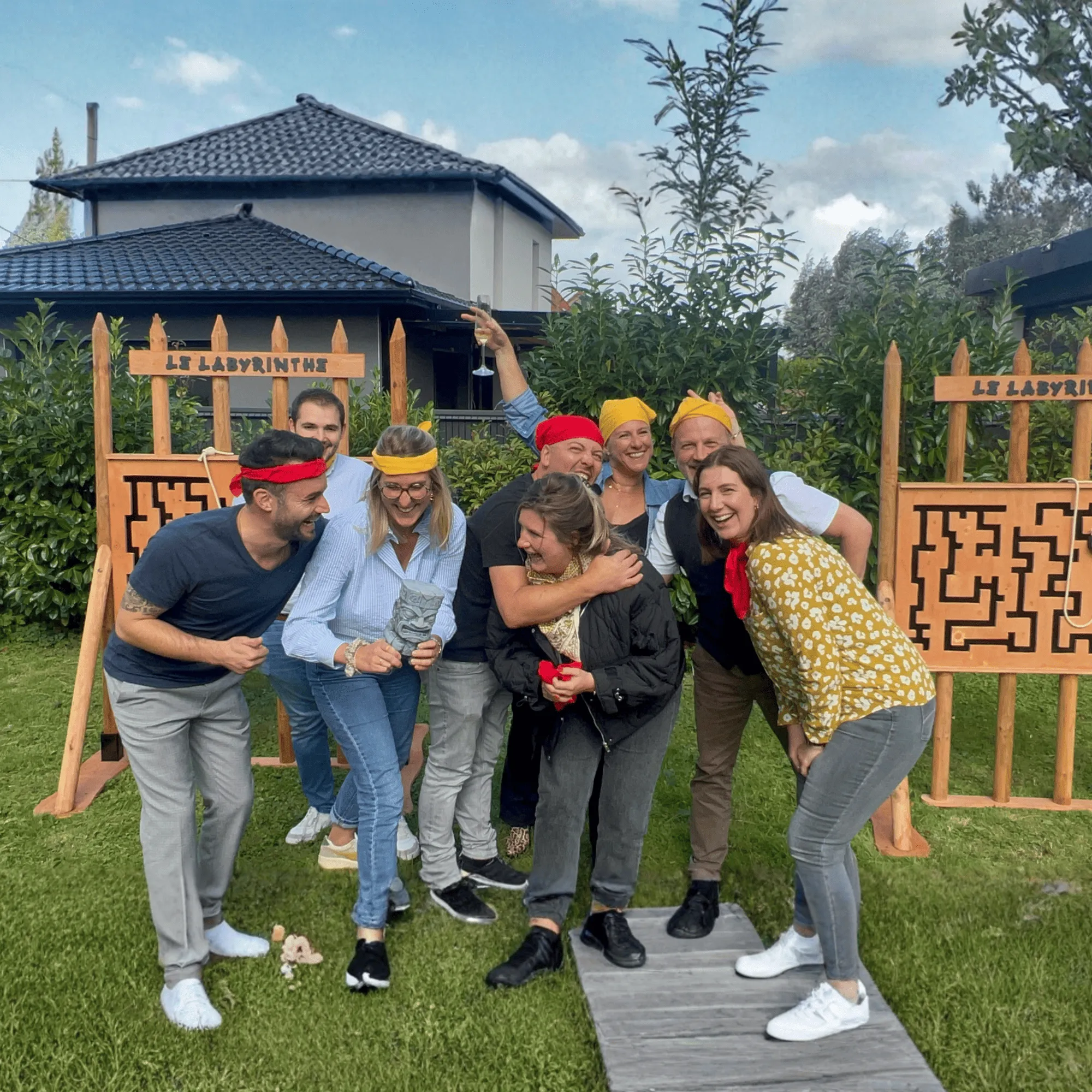 Un groupe de huit personnes en activité de team building en plein air, portant des bandeaux colorés et souriant devant des panneaux de labyrinthe en bois.