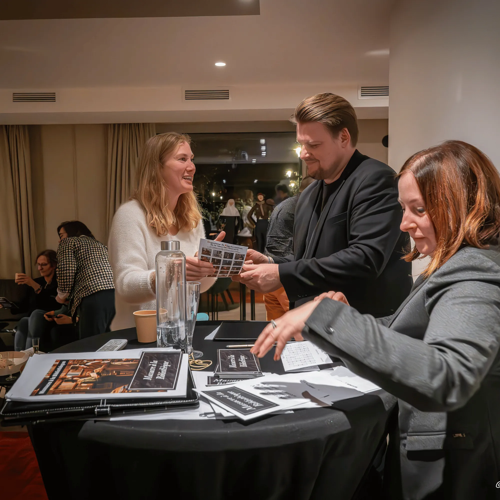 Trois personnes debout autour d'une table ronde noire, échangeant des documents dans un cadre intérieur chaleureux.