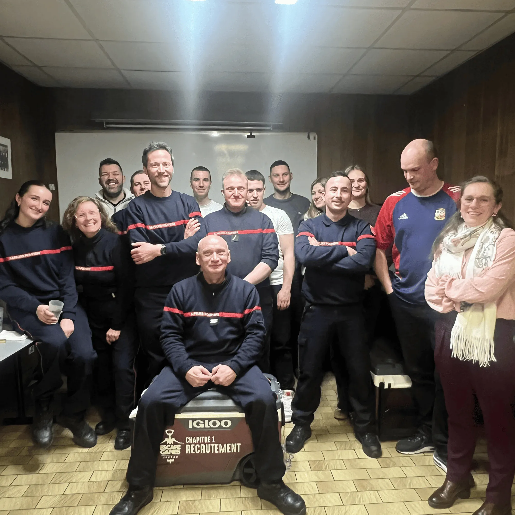 Un groupe de pompiers en uniforme posant dans une salle avec un homme assis sur une glacière marquée 'Chapitre 1 Recrutement'.