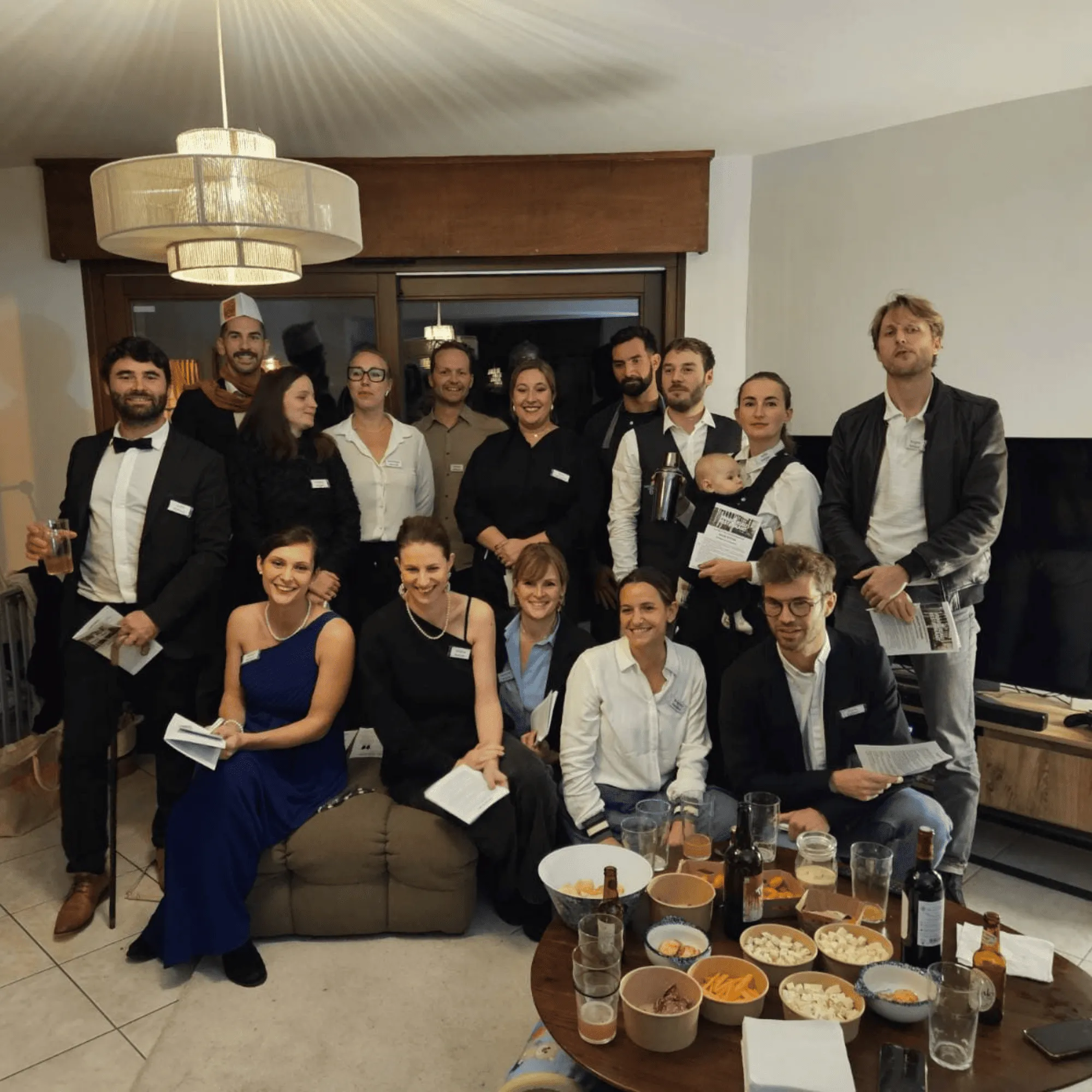 Groupe de personnes habillées pour une soirée, posant dans un salon avec une table garnie de boissons et snacks. Murder party