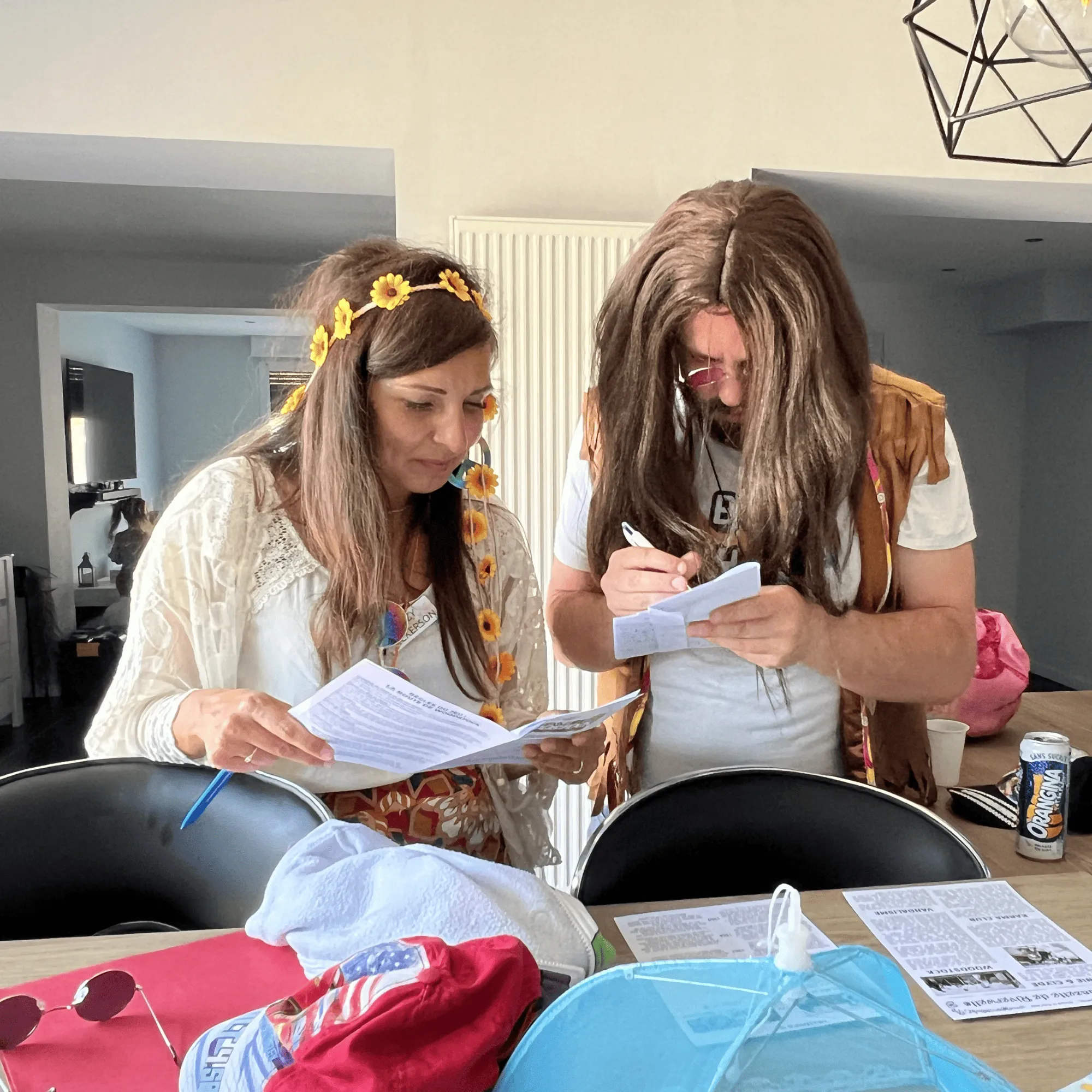 Deux adultes déguisés des années 70, une femme avec une robe à fleurs et un homme avec une perruque longue et des lunettes, lisent et écrivent sur des feuilles à une table.