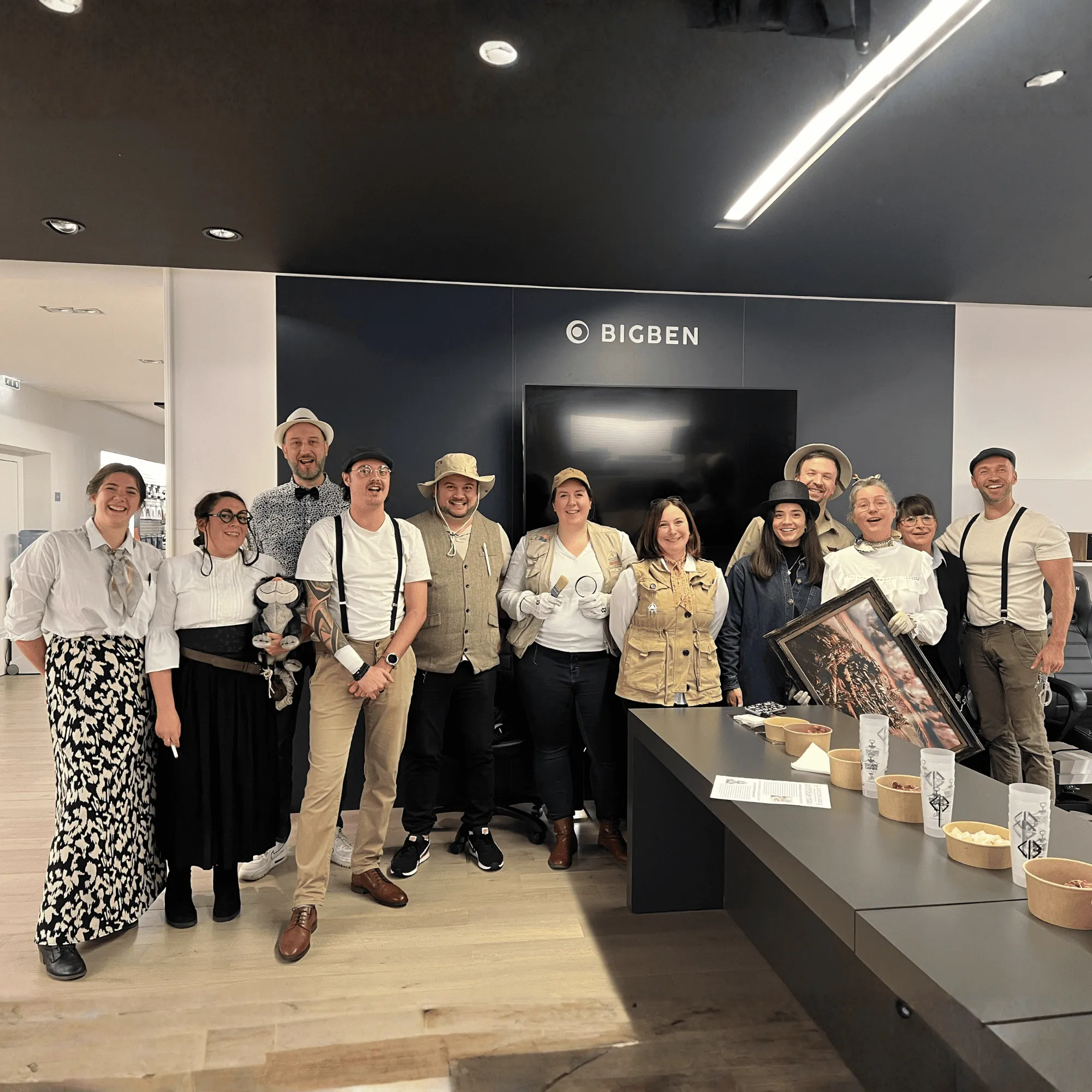 Groupe de personnes en costumes d'explorateurs et détectives posant devant un mur avec logo Bigben, à côté d'une table avec des bols de snacks et verres.