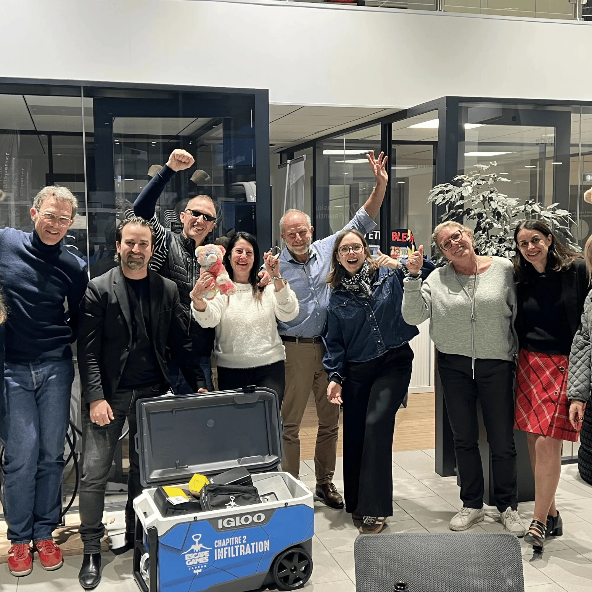 Groupe de personnes souriantes célébrant autour d'une glacière bleue marquée Escape Game Chapitre 2 Infiltration dans un espace intérieur moderne. detective party