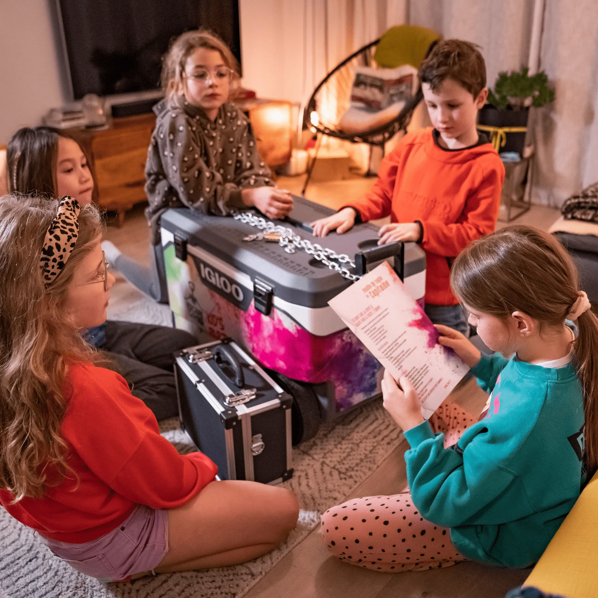 Cinq enfants assis en cercle dans une pièce, tentant d’ouvrir une grande glacière verrouillée par une chaîne et un cadenas.