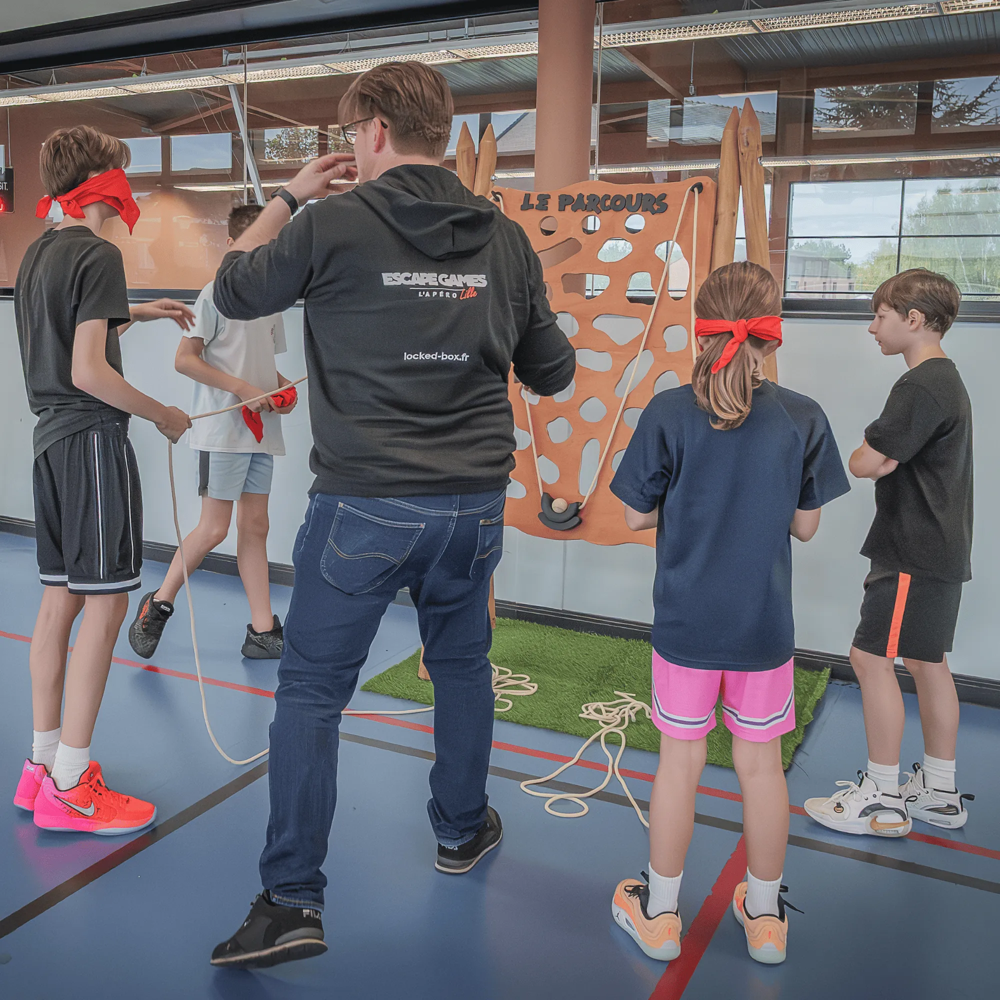 Un groupe d'enfants et un animateur jouent à un jeu d'adresse avec une planche en bois intitulée « Le parcours » dans une salle de sport.