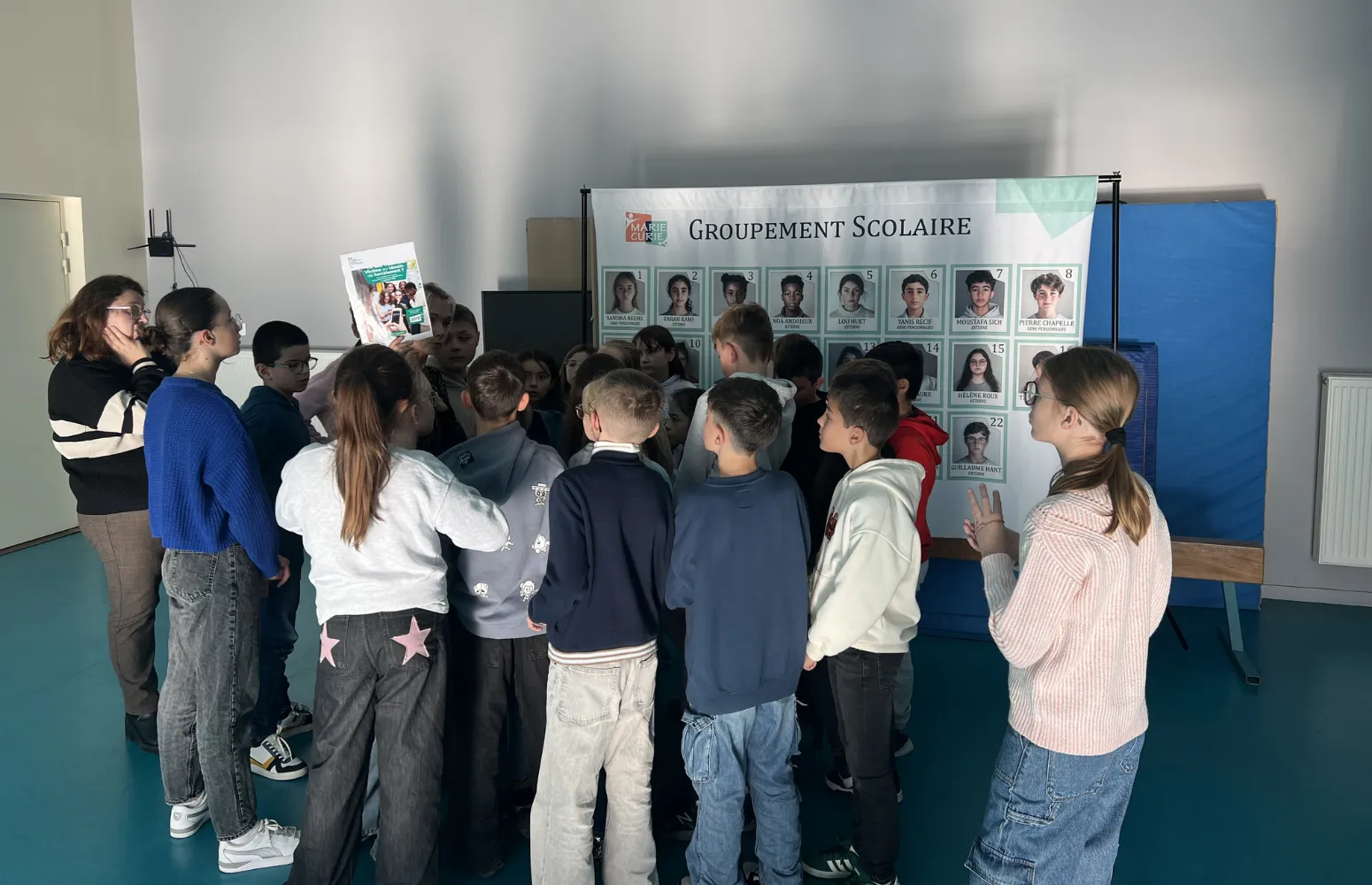 Groupe d'enfants regardant un grand panneau avec des photos et des noms d'élèves intitulé 'Groupement Scolaire'.