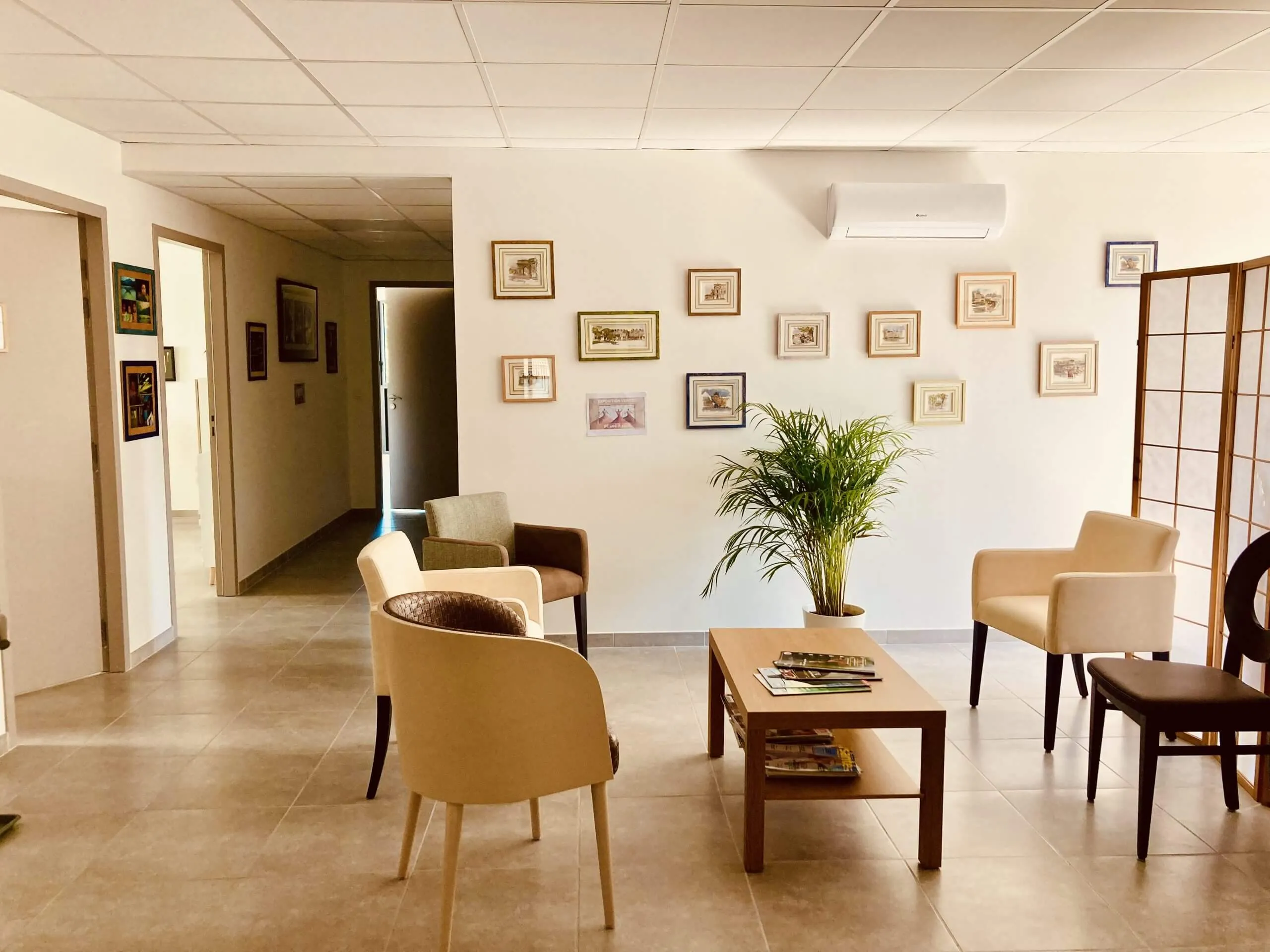 Waiting room with beige and brown chairs around a wooden coffee table, a potted plant, and framed pictures on a white wall with an air conditioner.