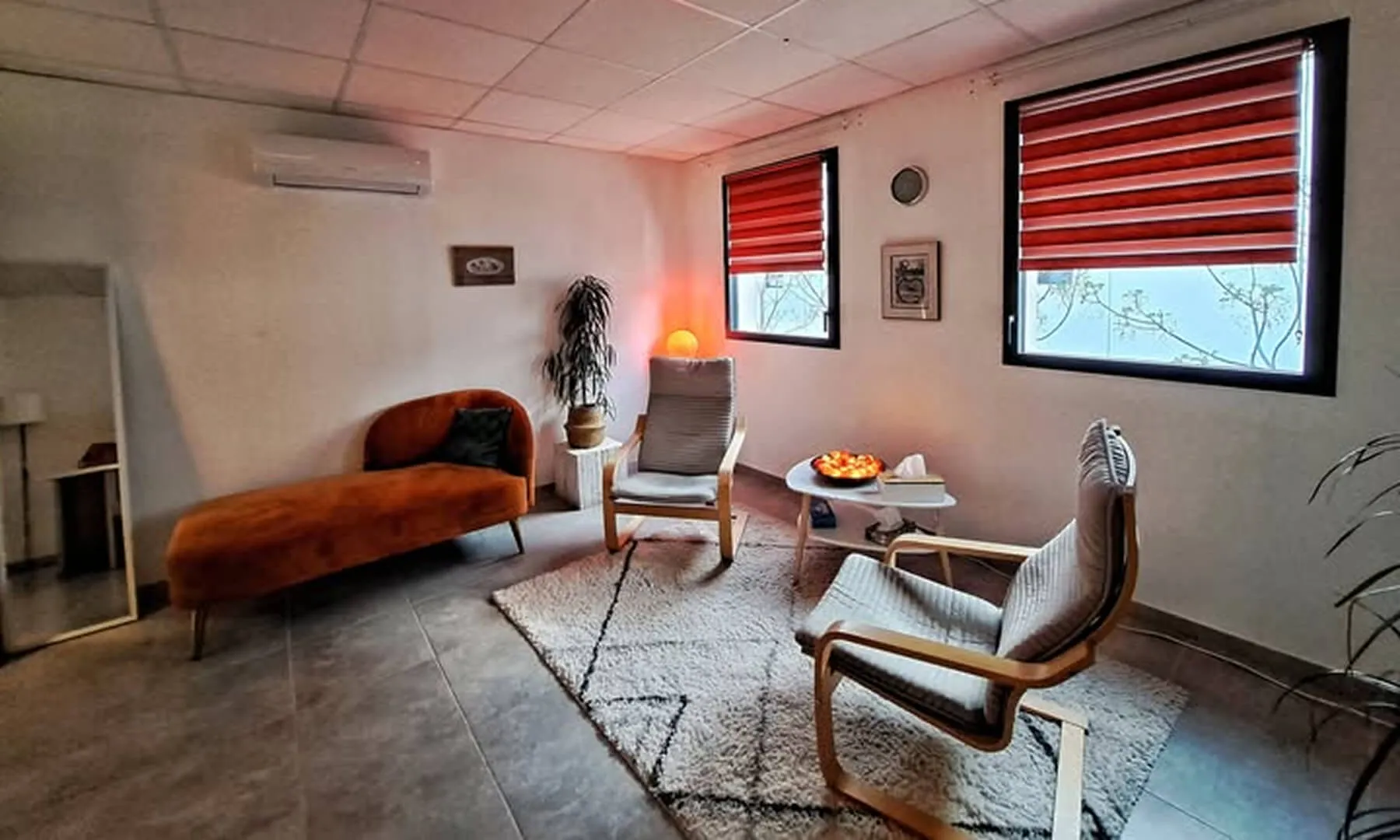 Cozy living room with two wooden armchairs with gray cushions, a rust-colored chaise lounge, red window blinds, and a white patterned rug.