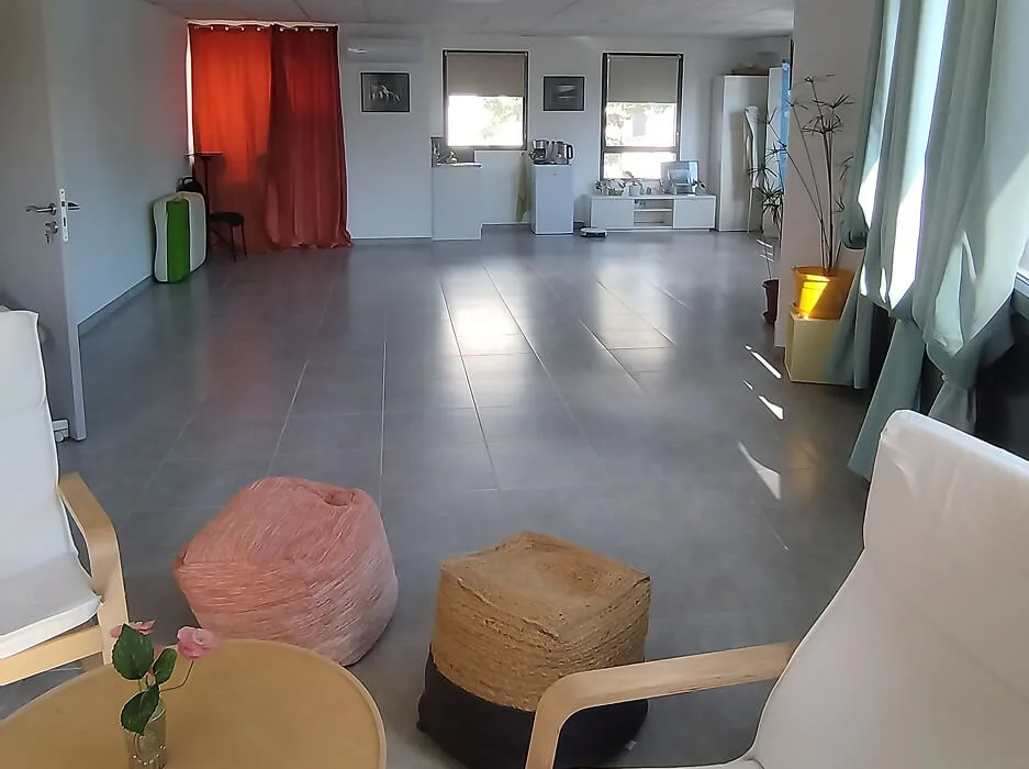 Spacious room with gray tiled floor, two white armchairs, a round wooden table with a small flower vase, two poufs, and windows with curtains letting in daylight.