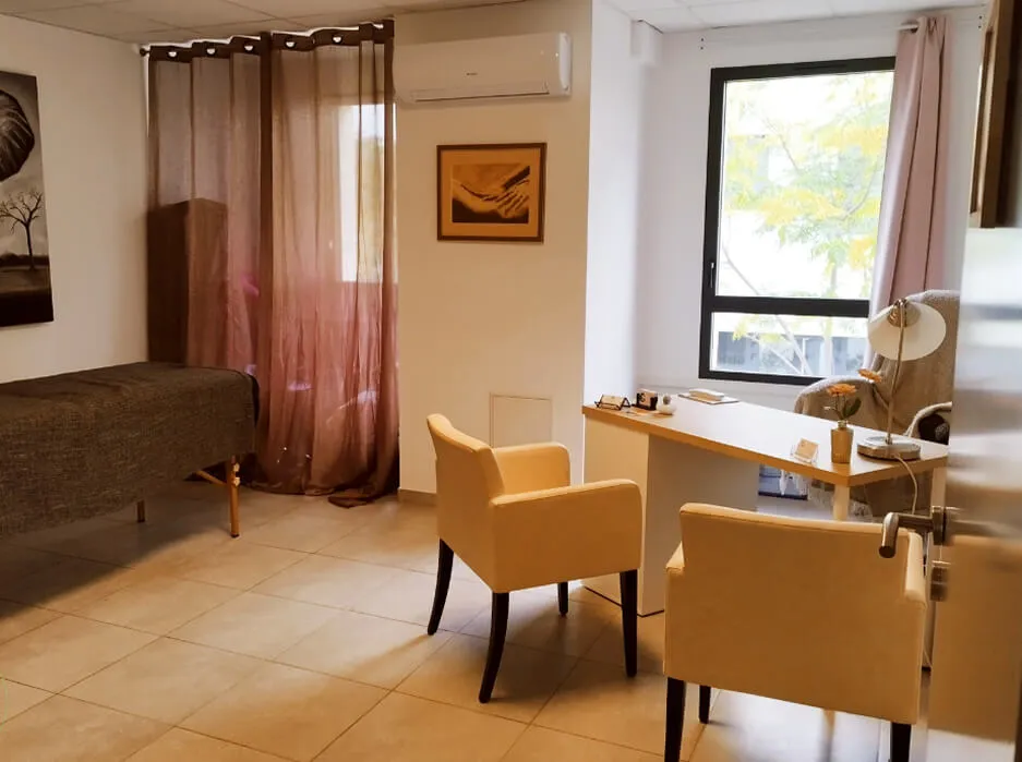 Cozy office room with a wooden desk, two beige chairs, a window with curtains, and a brown bed with a blanket.