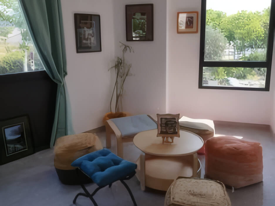 Cozy sitting area with a round wooden table, various cushioned stools and a window showing green foliage outside.