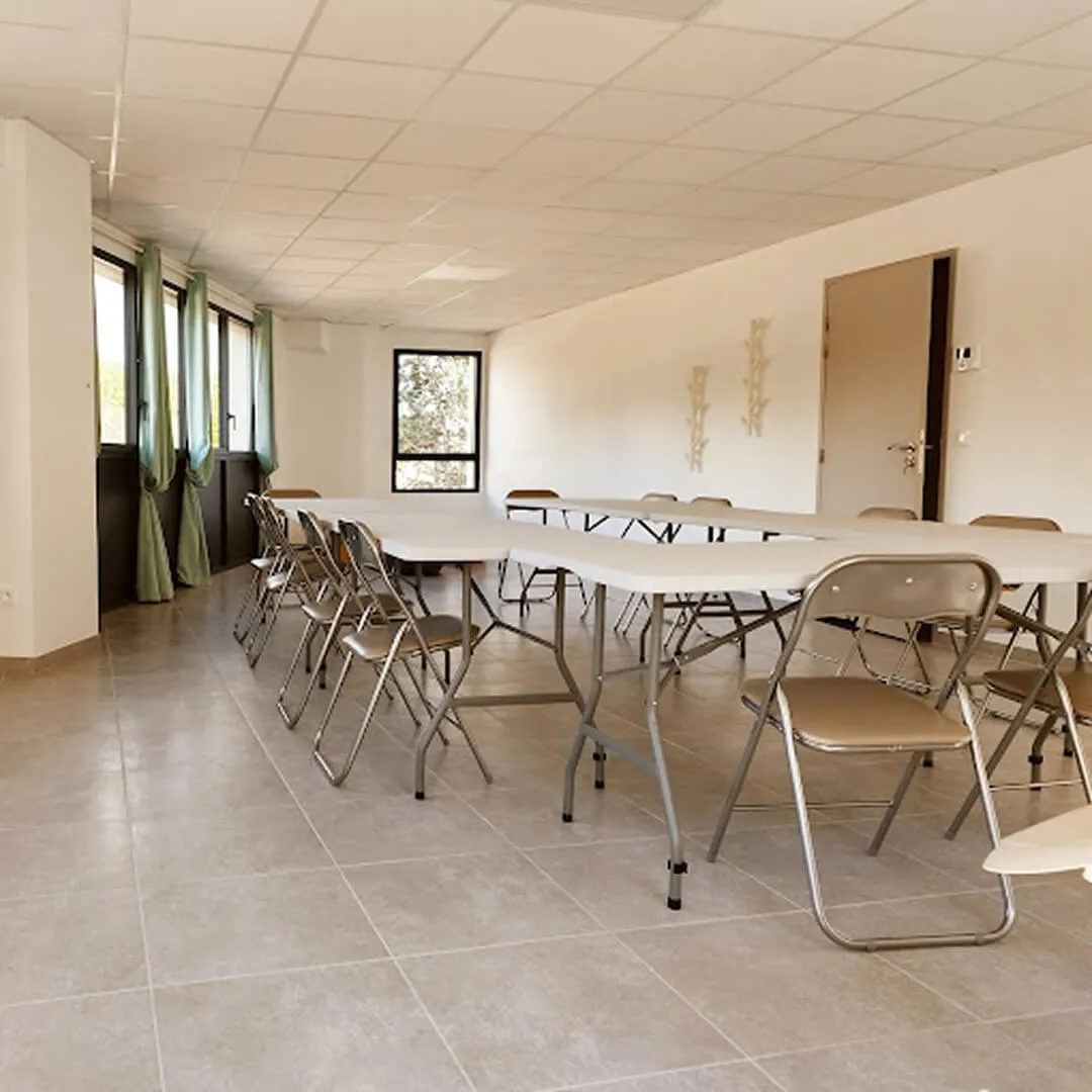Bright conference room with folding tables arranged in a U-shape and metal folding chairs, large windows with green curtains.
