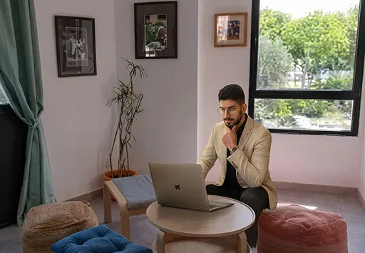 Man in beige blazer working on a laptop seated at a round wooden table in a cozy room with framed pictures and greenery outside the window.
