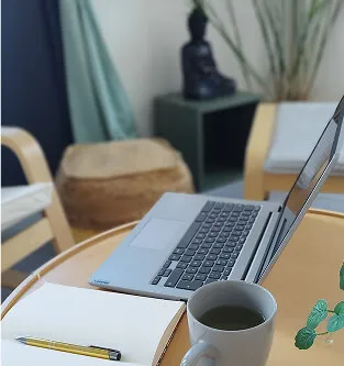 Workspace with an open laptop, a white coffee mug, an open notebook, and a pencil on a round wooden table.