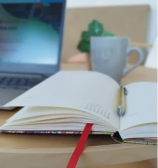 Open notebook with a red ribbon marker and pen resting on a wooden table, with a laptop and a gray mug in the background.