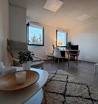 Modern office room with a wooden chair, white desk, plants, rug, and two windows letting in natural light.