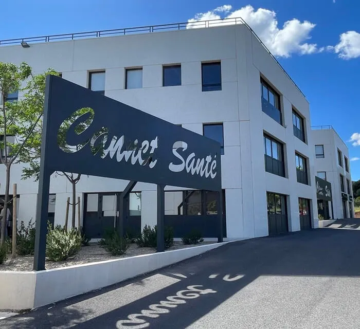 Modern white three-story building with large windows under a blue sky, featuring a black metal sign with the words 'Pole Santé' casting a shadow on the ground.