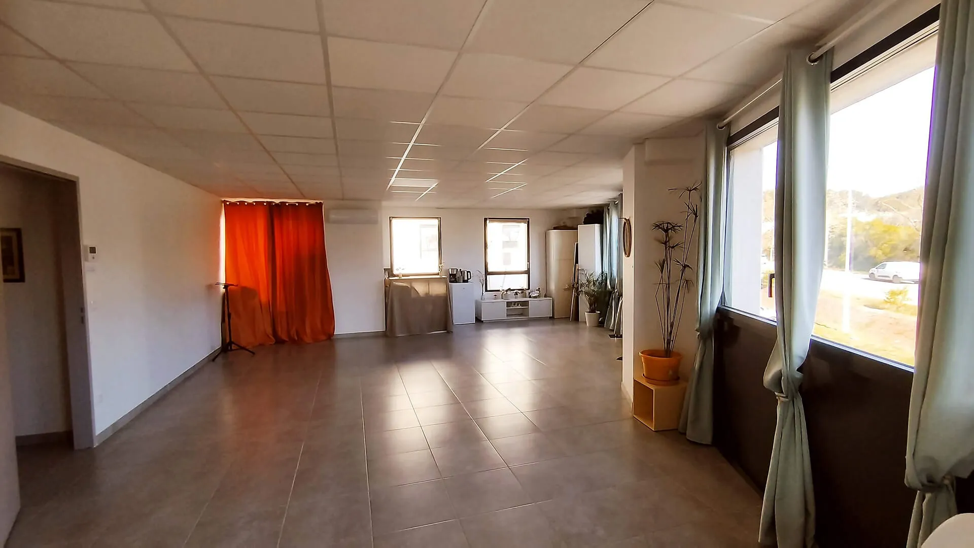Spacious room with tiled floor, large windows with light curtains, an orange curtain, and minimal furniture including plants and a small cabinet.