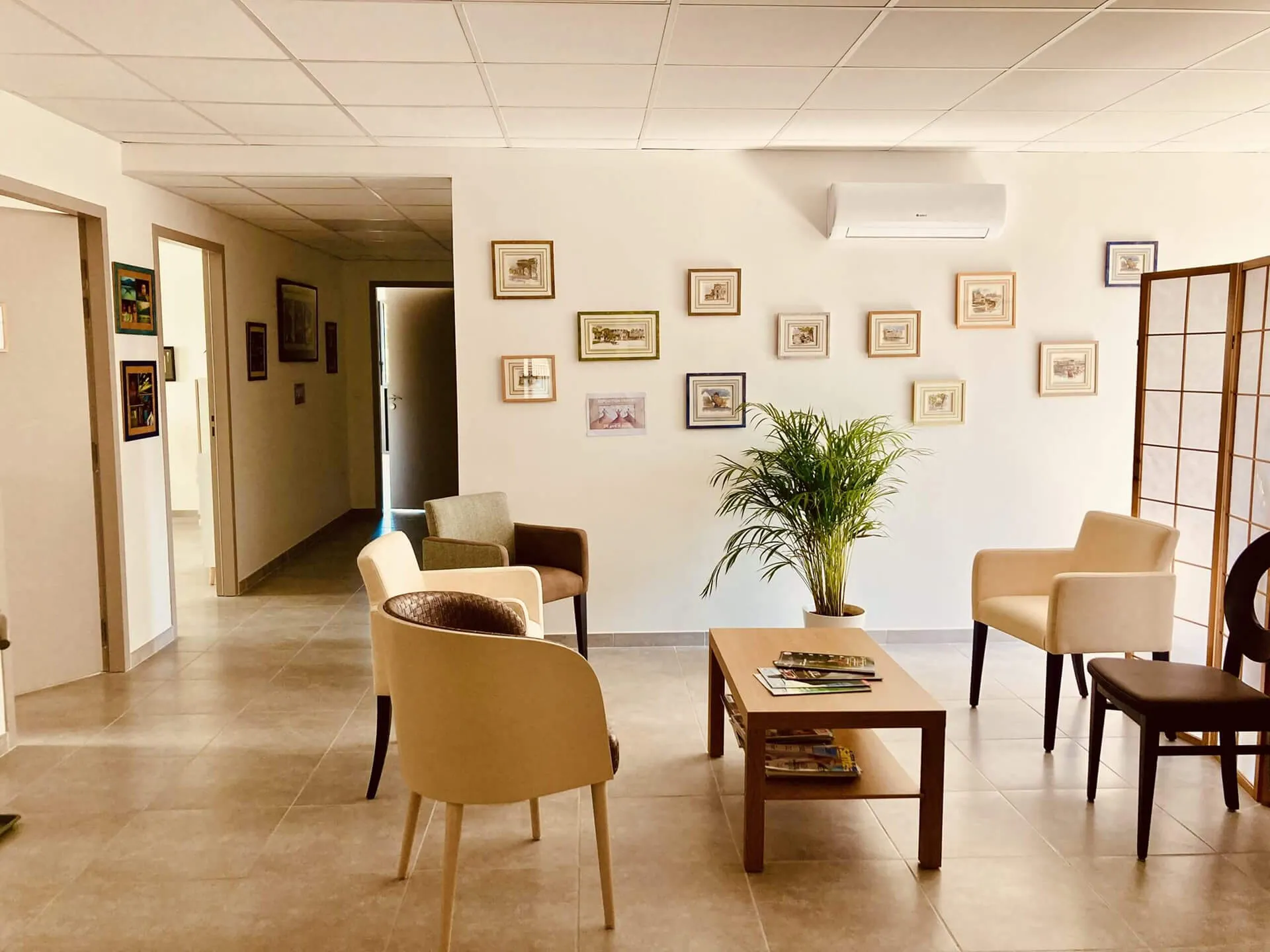 Modern waiting room with beige and brown chairs around a wooden coffee table, a potted plant, framed pictures on the wall, and a split air conditioner.