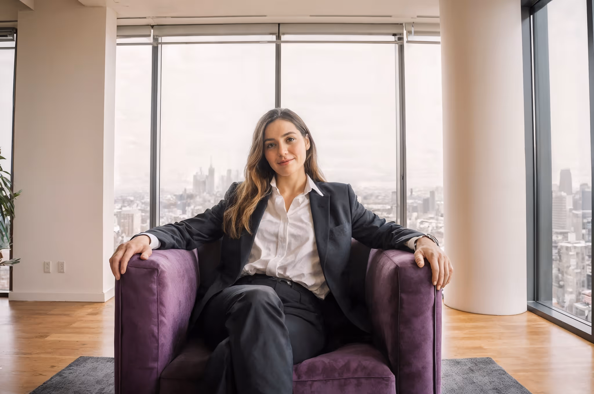 Confident woman in business suit sitting on purple armchair in modern office with city view.