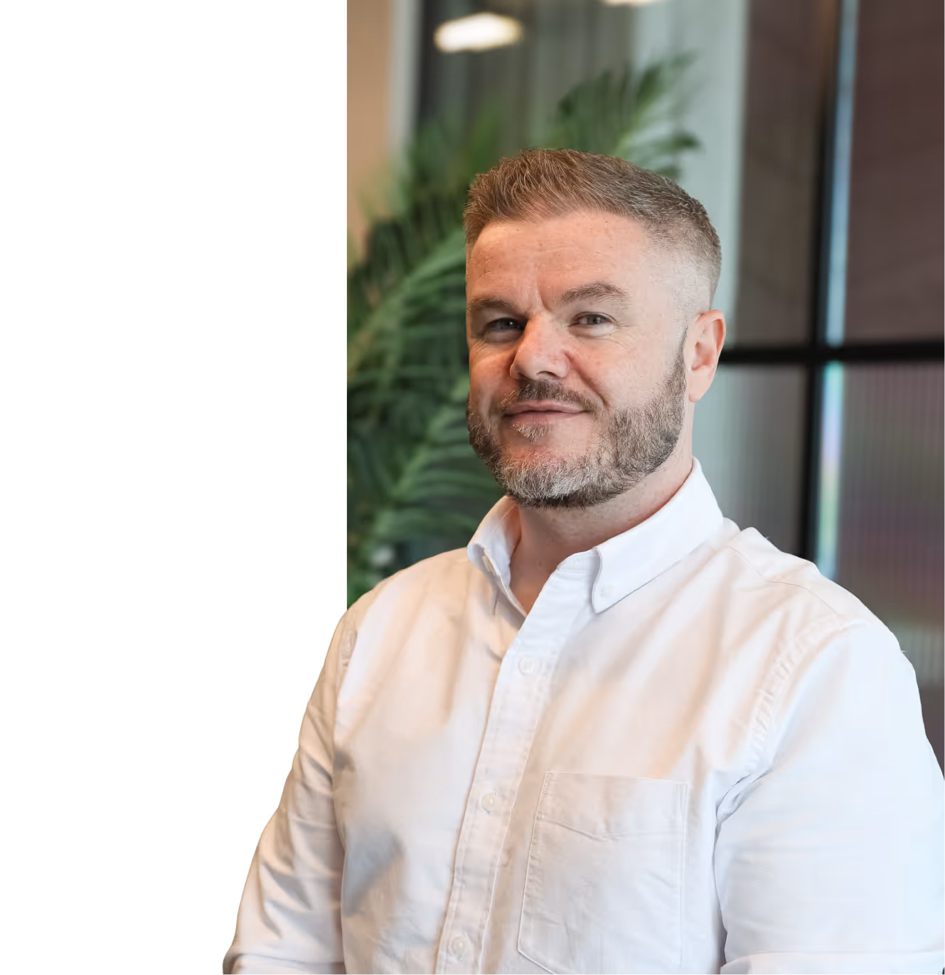 Man with short hair and beard wearing a white button-up shirt, standing indoors with blurred plant and window in the background.