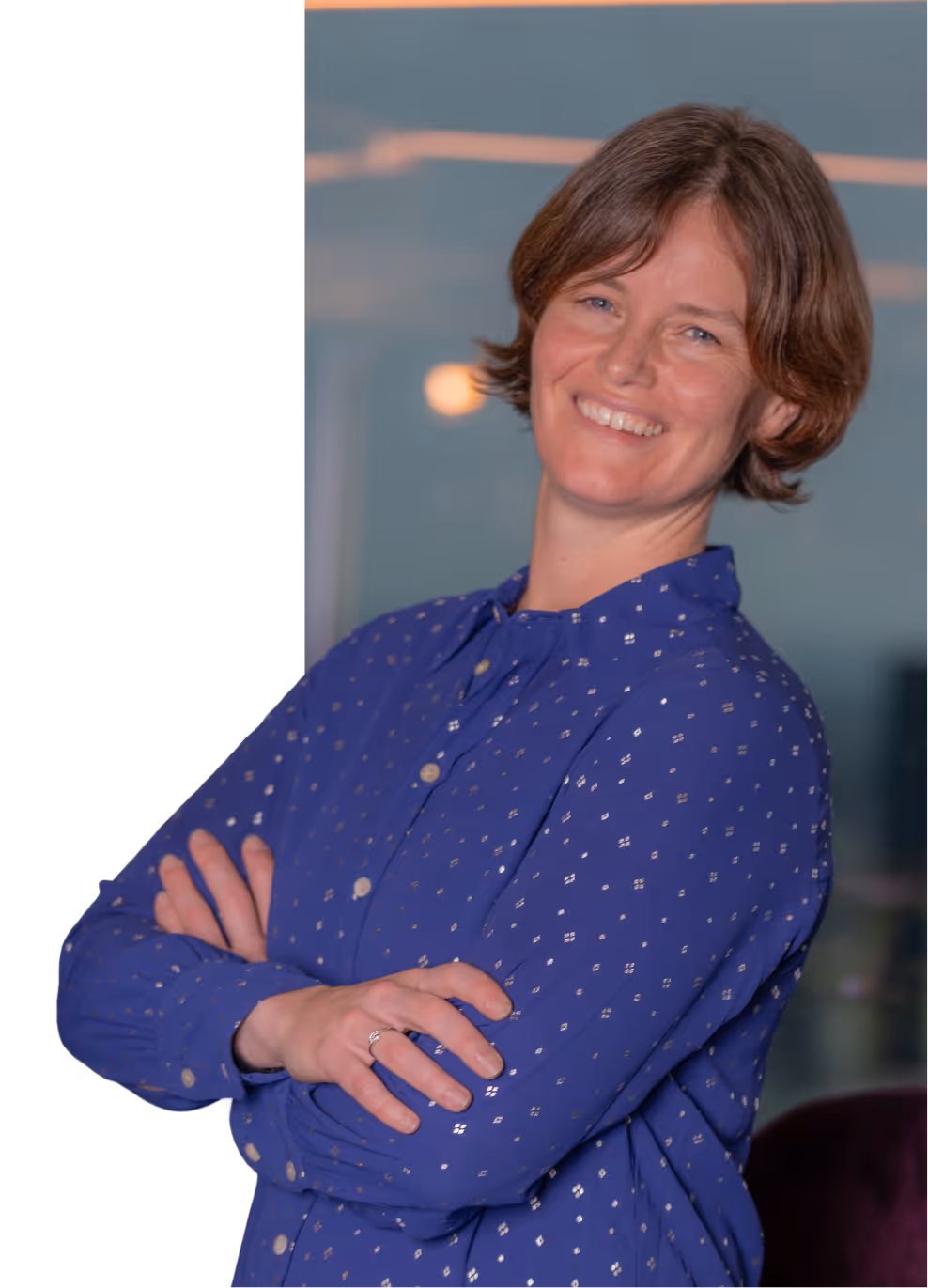 Smiling woman with short brown hair wearing a blue patterned blouse standing with arms crossed.