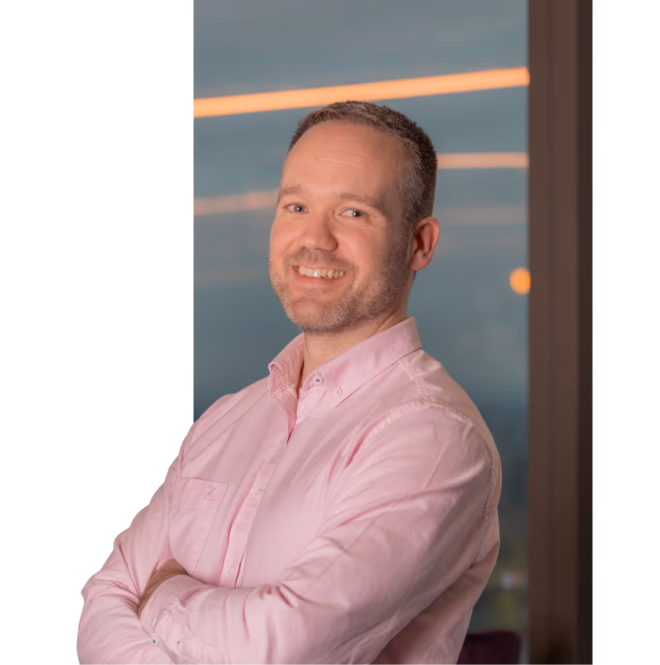 Man with short hair and beard wearing a light pink button-down shirt, smiling with arms crossed.