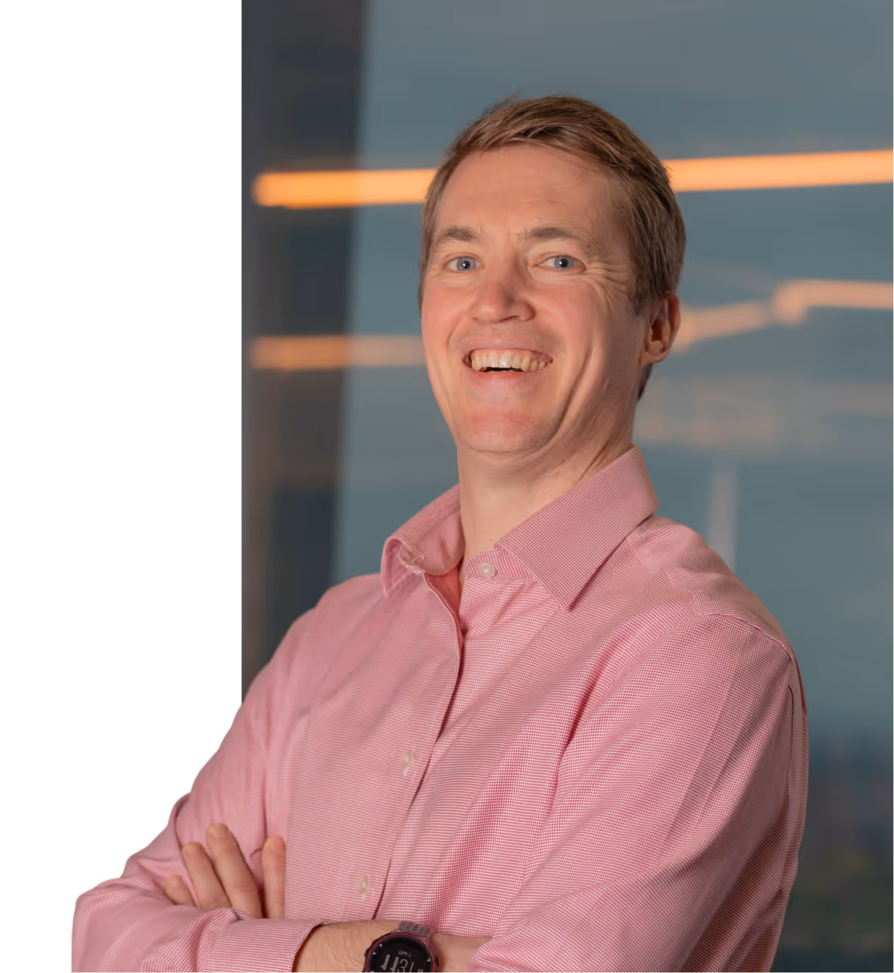 Smiling man wearing a pink button-up shirt and smartwatch, standing with arms crossed.