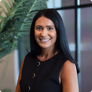 Smiling woman with long black hair wearing a sleeveless black top, standing indoors with a plant and window in the background.
