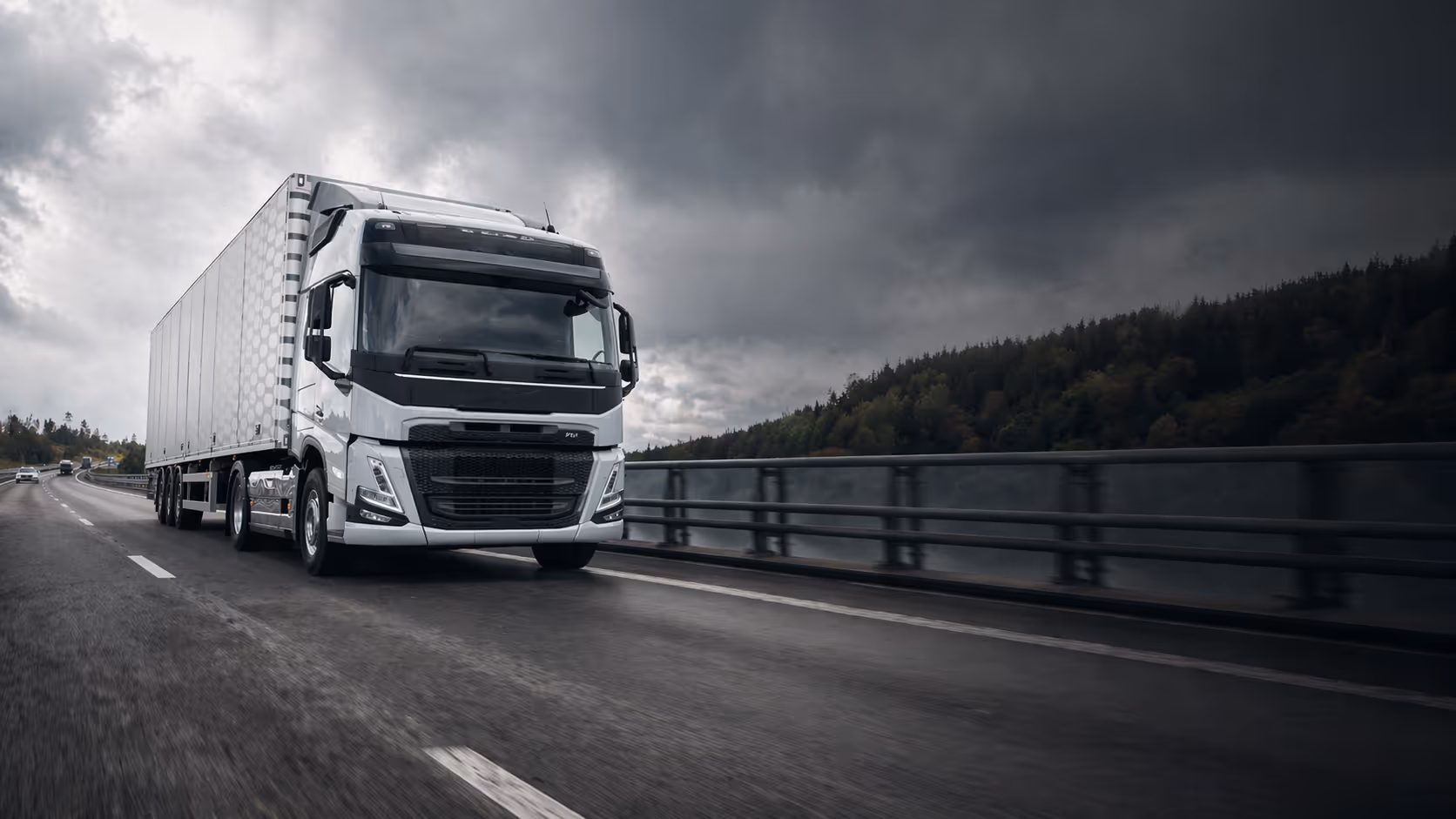 White semi-truck driving on a highway under a cloudy sky with trees in the background.