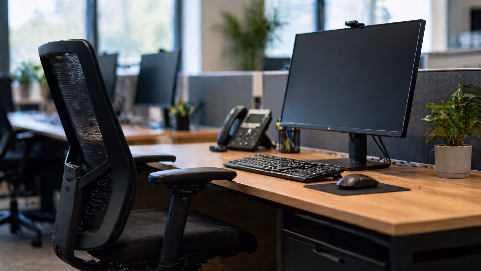 Modern office workstation with a black mesh chair, computer monitor, keyboard, mouse, desk phone, and a small potted plant on a wooden desk.